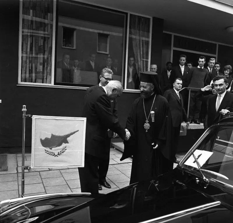 President Makarios of Cyprus, in clerical robes, beside a stand bearing his country's flag during a state visit to West Germany in the 1960s